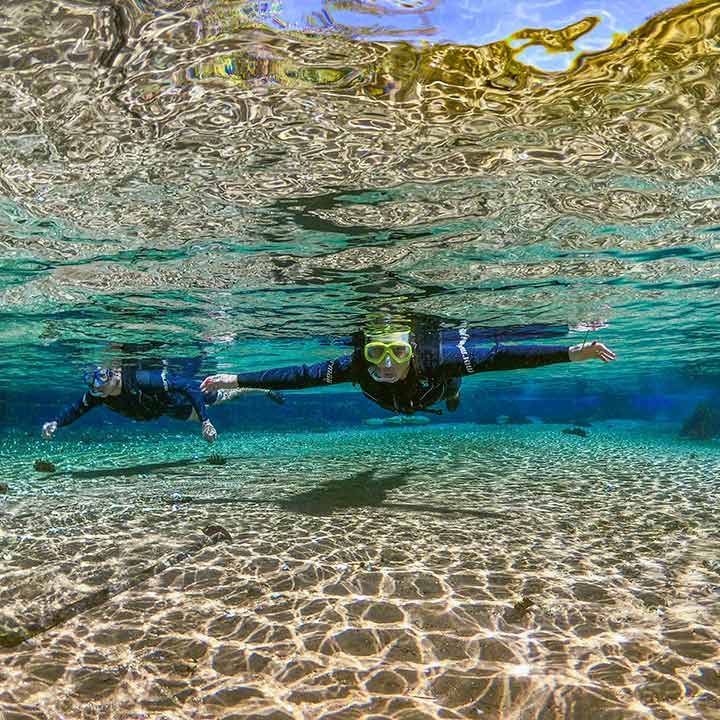 Pessoa fazendo mergulho com snorkel em águas cristalinas, com fundo de areia e pedras, sob a superfície da água com reflexos dourados e azulados.