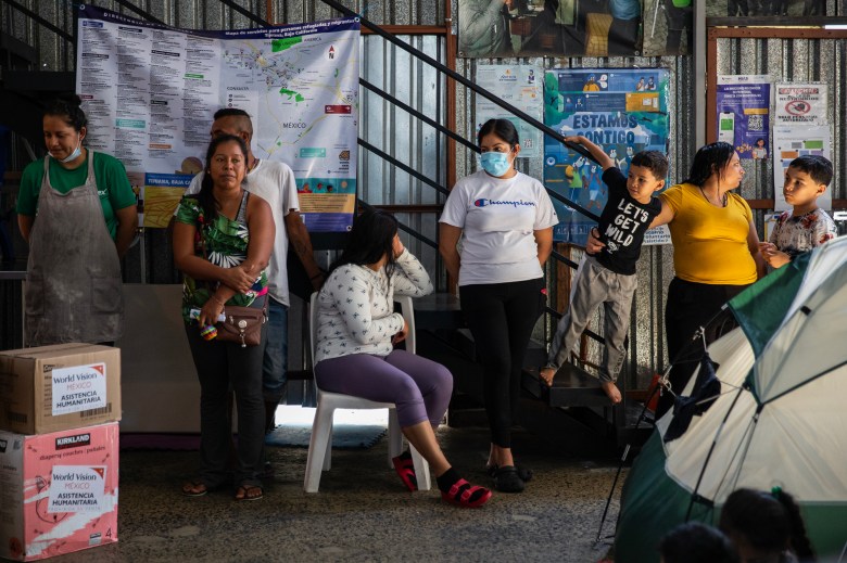 A group of people, including women and children, stand and sit inside a shelter facility. Some are leaning against a wall or stair railing, with maps and informational posters displayed behind them. One woman wears a face mask, and another sits in a plastic chair, resting her head on her hand. Boxes labeled with 