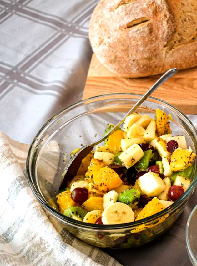 A glass bowl of fruit salad with oranges, bananas, grapes, kiwi, and chia seeds, next to a loaf of bread.