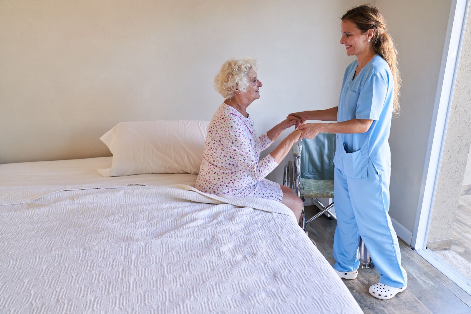 A caring nurse helps an older adult woman get up from bed and into a wheelchair.