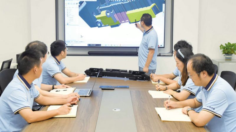 Alguém apresenta um projeto técnico em uma tela para um grupo em uma mesa de conferência, enquanto eles tomam notas.