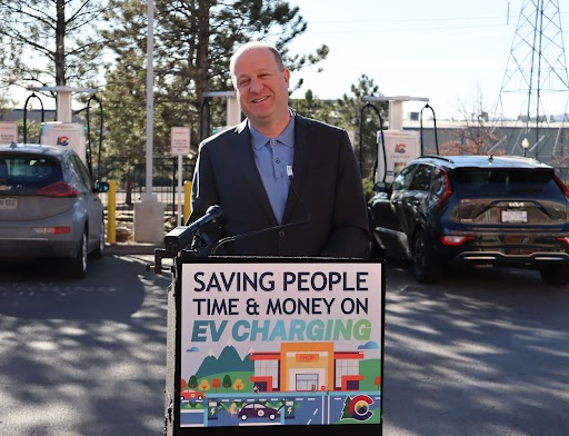 Governor Polis stands behind a podium that says saving people time and money on EV charging. There are EVs plugged in charging at a charging station in the background.