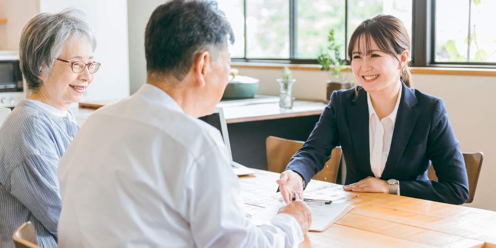  年配夫婦と女性担当者がリビングのテーブルで売却相談を行い笑顔で会話する場面