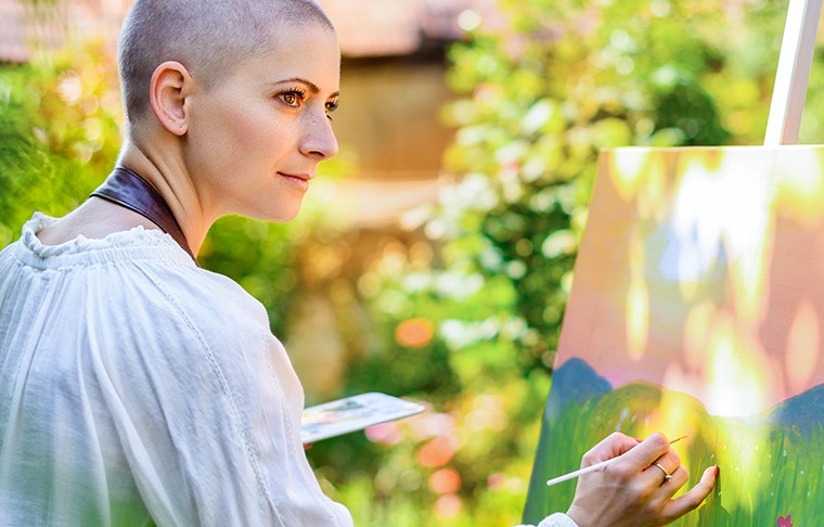 Mulher jovem e bonita relaxando enquanto pinta uma tela de arte ao ar livre em seu jardim. Sobrevivente de câncer, atenção plena, terapia de arte, conceito de criatividade.