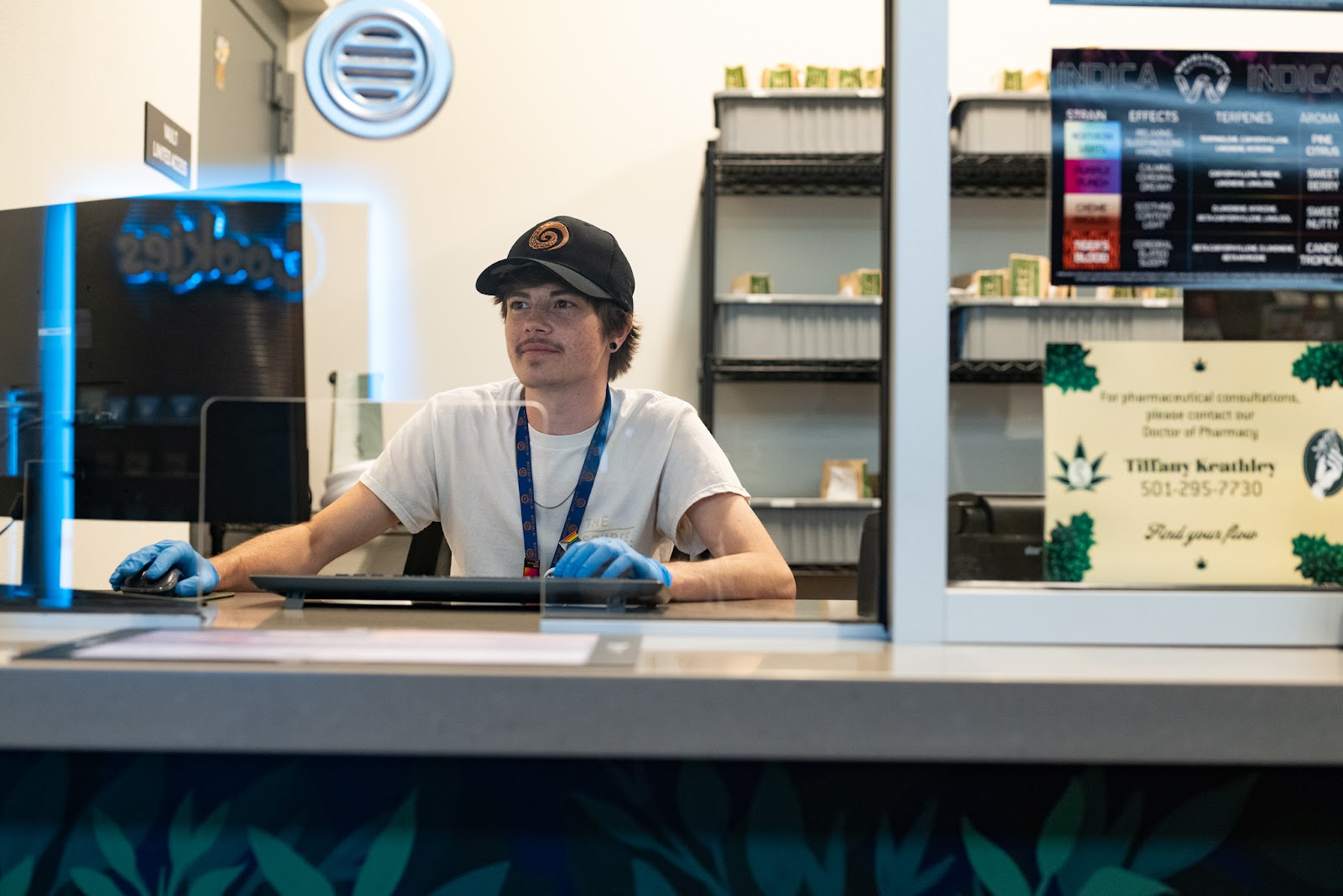 A young budtender, Eli Hanna, sits behind a glass wall working on a computer wearing blue rubber gloves. The phone number for pharmaceutical consultations on medical marijuana is posted on the glass alongside a menu for Wavelength Extracts premium vape cartridges. 