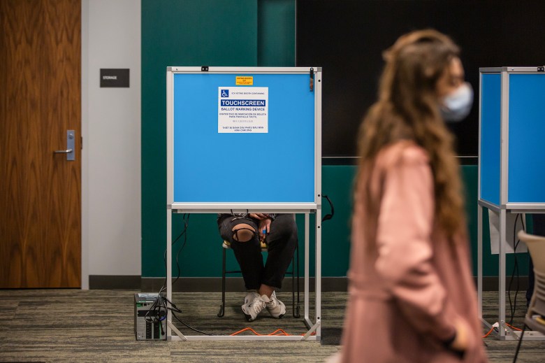 A person is seated in a wheelchair-accessible voting booth with a blue privacy screen, their legs and hands visible as they hold a pen. In the foreground, another individual, wearing a mask and blurred in motion, walks by, partially obscuring the booth. The setup includes a touchscreen ballot marking device for accessibility, and a storage room door is visible in the background.