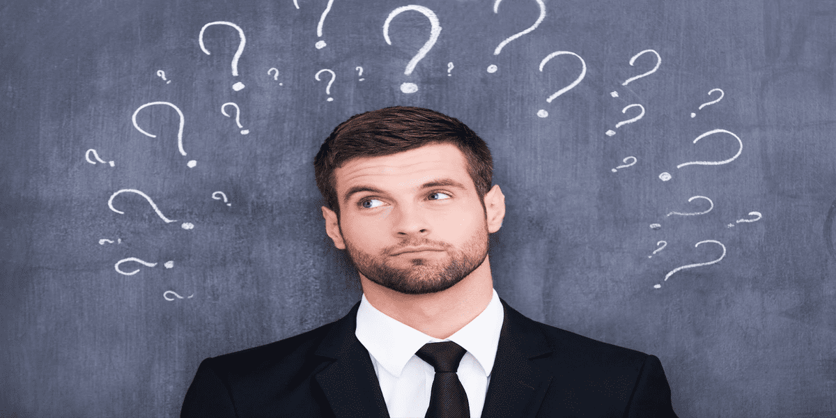 Young man in a suit looking puzzled with multiple question marks drawn on a chalkboard behind him