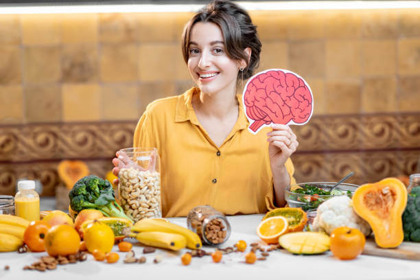 Smiling woman holding an illustrated brain image while standing behind a table filled with fresh vegetables, fruits, and nuts, representing brain-boosting foods.