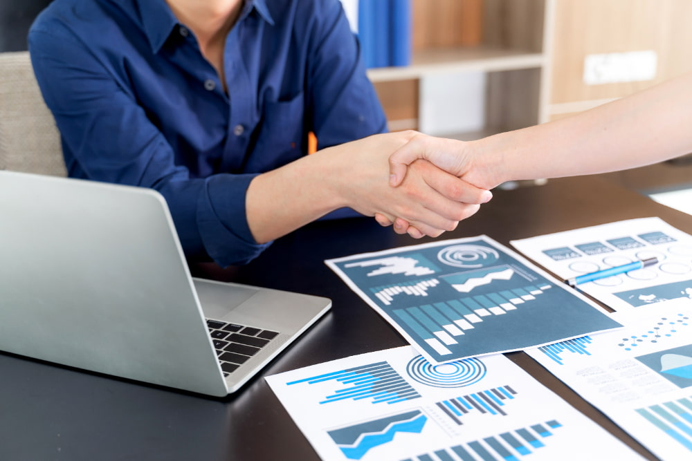 Two individuals shaking hands over a desk with charts and an open laptop.