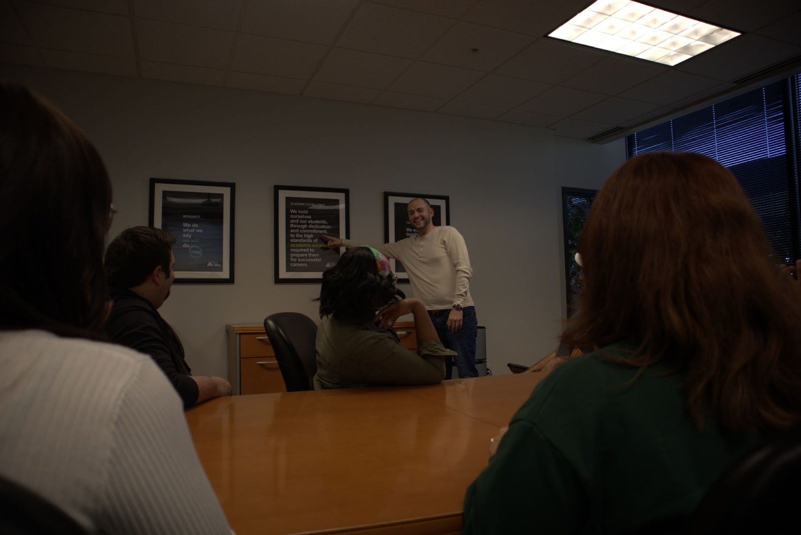 A group of people gathered in a conference room, sitting around a wooden table. One individual, standing near the wall, is smiling and appears to be presenting or speaking, while others are seated and clapping. The wall displays framed posters highlighting values like accountability, integrity, and academic excellence, creating a professional and motivational atmosphere.