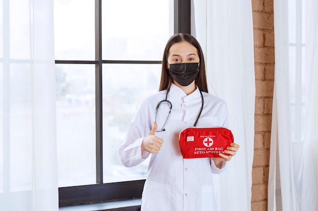 Doctor with stethoscope and black mask holding a red first aid kit and showing thumb up. 