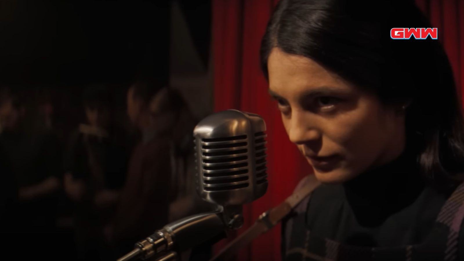 Joan Baez with long dark hair speaks into microphone in a dimly lit venue.