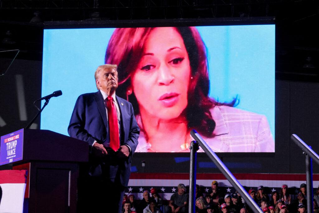 Republican presidential nominee and former US President Donald Trump looks on as Democratic presidential nominee and U.S. Vice President Kamala Harris' face appears as a video plays on a screen, during a rally at Huntington Place in Detroit, Michigan, US, 18 October 2024 (Photo: Reuters/Brian Snyder).