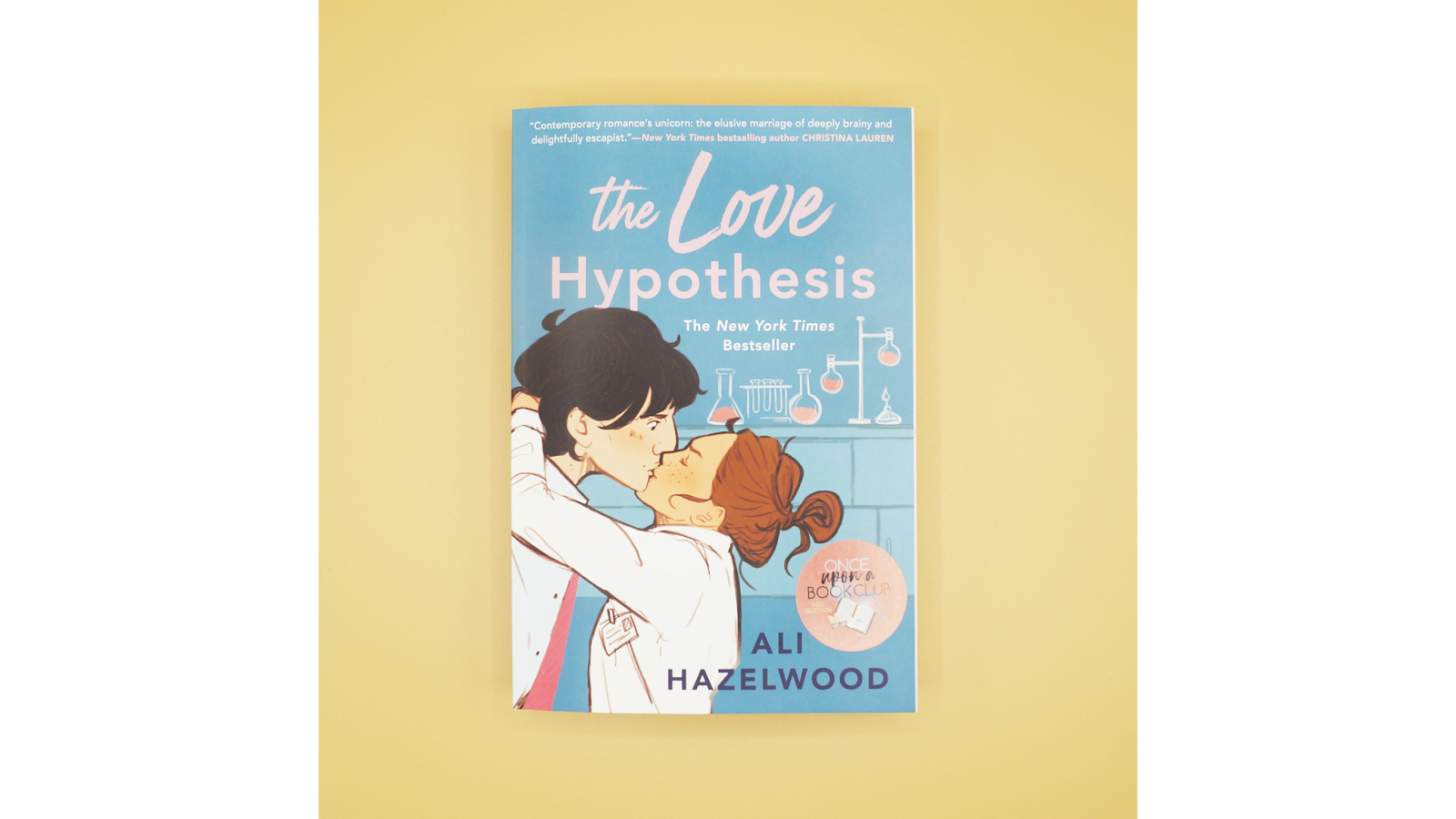 The cover of The Love Hypothesis by Ali Hazelwood, showing two characters kissing in a lab setting with a yellow background.