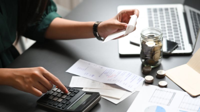 A person uses a calculator at a paper-strewn desk with coins and a notebook.