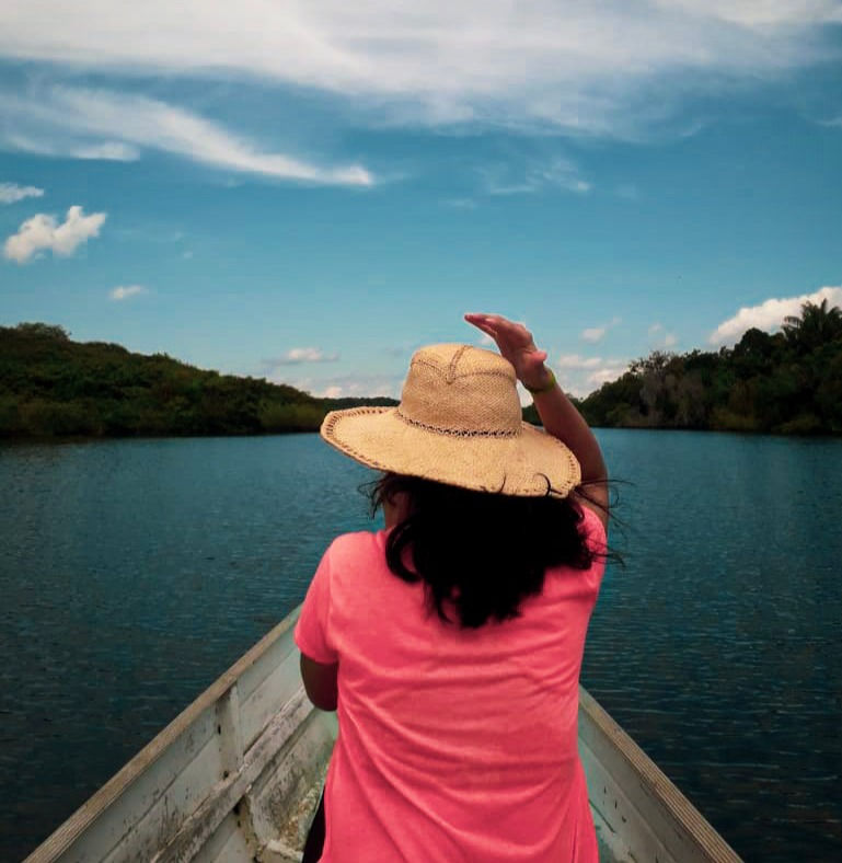 Mulher de costas na proa de uma canoa navegando um rio, ao fundo e nas laterais uma densa floresta a cerca.