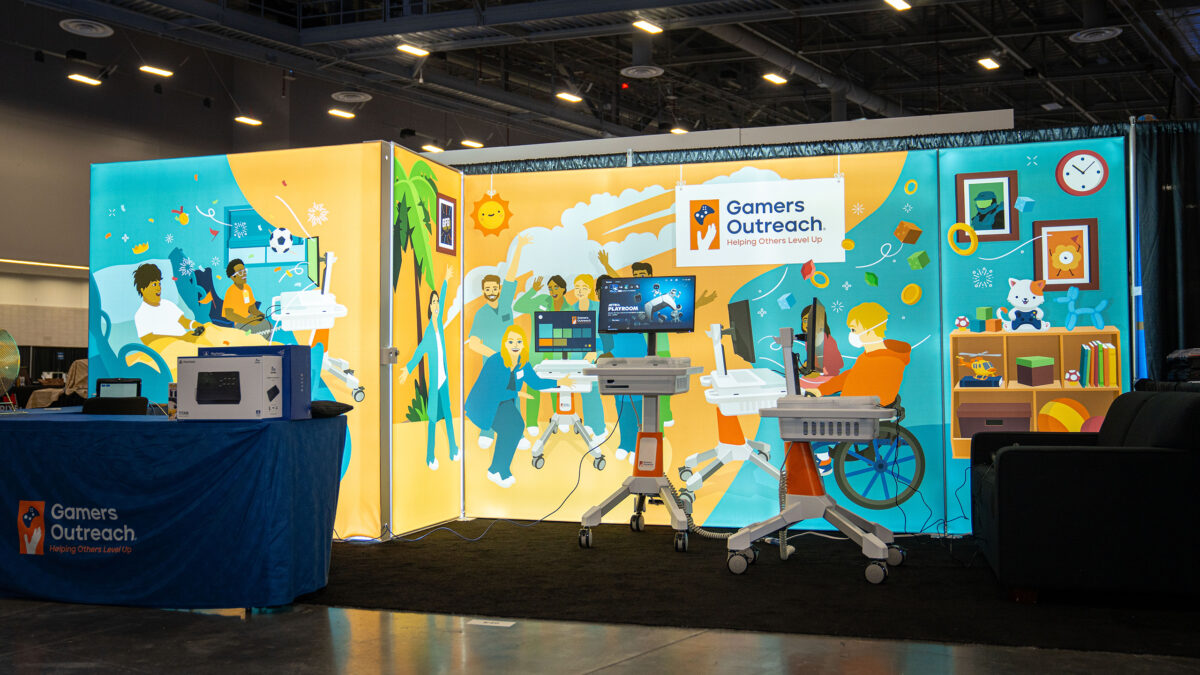 Wide view of the new Gamers Outreach booth, decorated in illustrations that were inspired by the GO Kart 3.0 hospital deliveries. Kids gaming, and hospital staff celebrating the GO Karts.