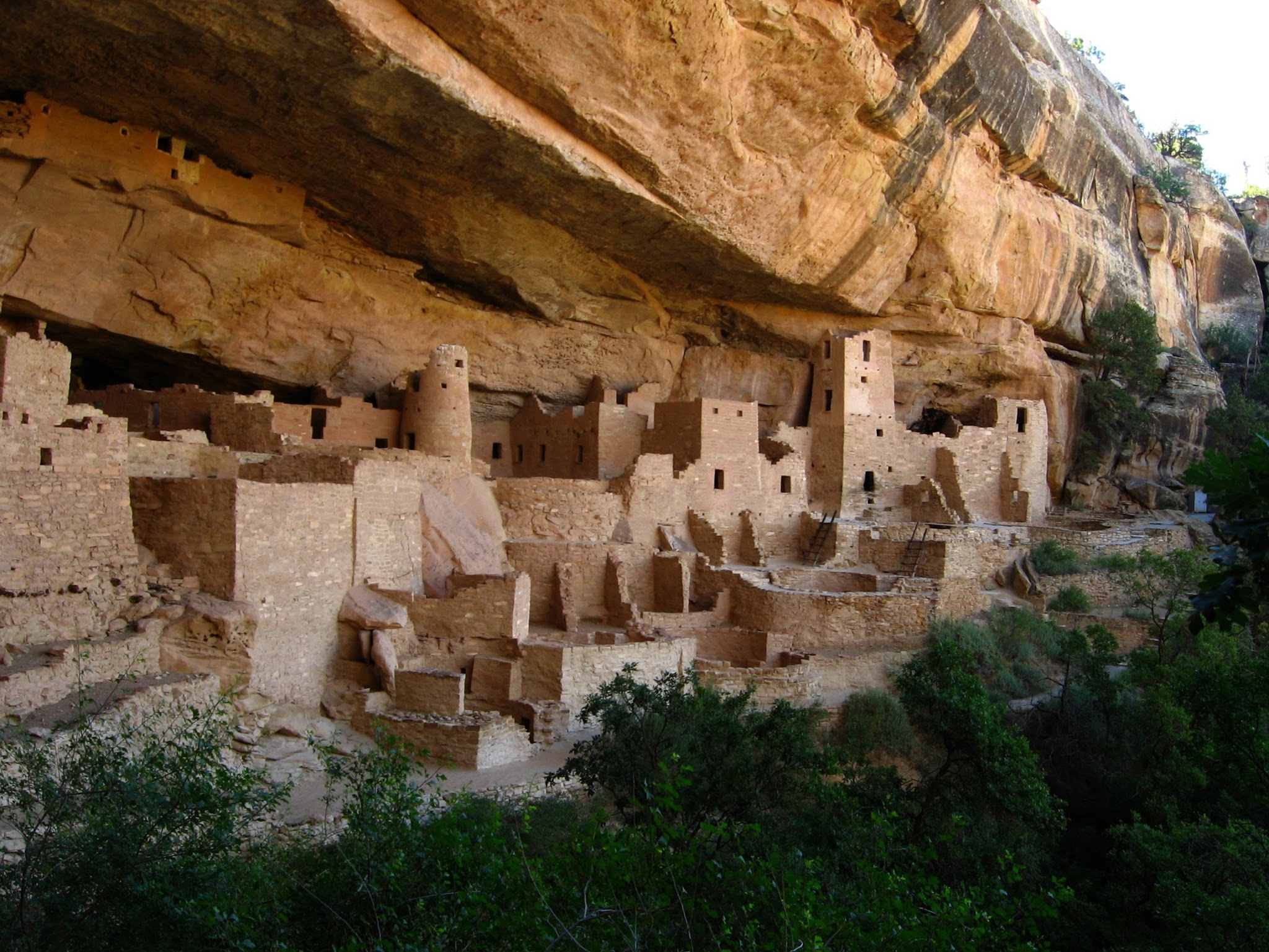 Architecture. Dwellings large enough for humans to inhabit are carved out of the living rock in cliff enclosures.
The site contains about 150 rooms built of stone and mortar. The cliffside ascends into a plateau area with copious greenery and vegetation. Beneath the dwellings, the valley below is lush and green in this image. The dwellings themselves, carved out into the side of the cliff, comprise multiple layers and stories with rugged stairs leading to each level.
