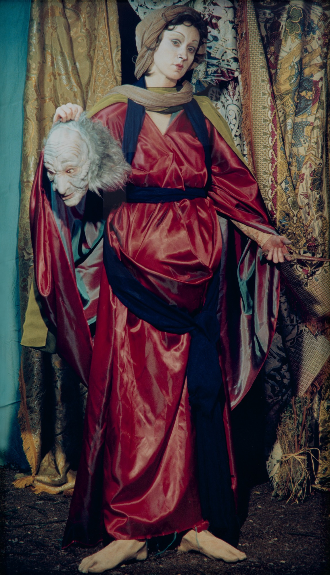 Photograph. A woman in an archaic, red dress holds scissors in her left hand, a ghoulish halloween mask in her right.
The photograph has been styled in such a way that it resembles a Baroque oil painting. Shiny, silky, patterned fabrics hang behind the full figure of the woman. Her wavy, brown hair is swept back under a shawl that wraps over her head and drapes across her shoulders. She stares out at the viewer calmly, holding the mask as if it is a decapitated man's head. Her abdomen is swollen as if she is in the late stages of pregnancy.

