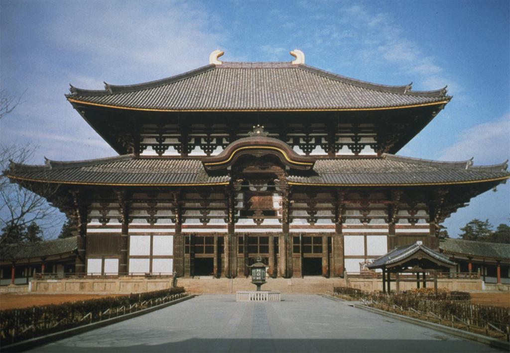 Architecture. Large building with a white surface, brown trim, three main stories, and two levels of curved roofs.
The three-story building is situated on a lush, green lawn with trees and bushes. Walls stretch out from either side, creating a barrier such that one could only enter from the main door in the center. Visitors approach the entrance from a path leading to the central door, their size offering perspective on the scale of the building. At its peak, the building is at least thirty times as tall as the average human. At the very top Are two horn-shaped features adorning the curved roof. The brown trim over the building forms a series of rectangular, triangular, and trapezoidal patterns.

