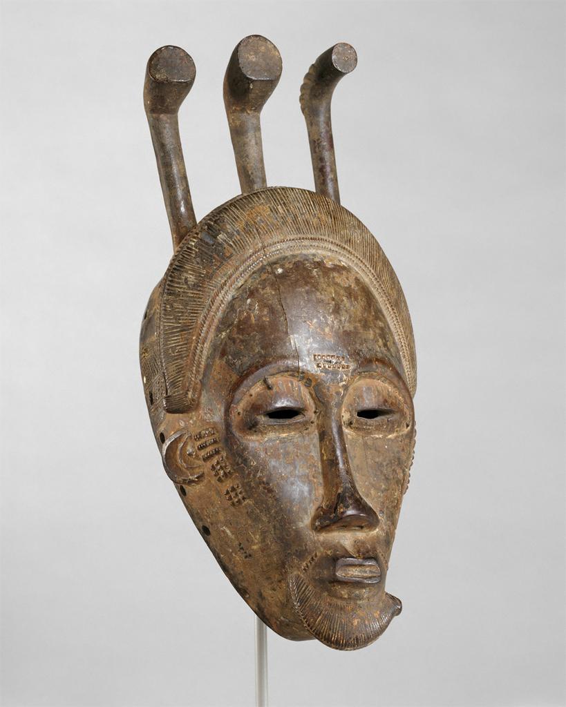 Mask. Carved wooded face with abstracted, geometric human features, with a long, straight nose and small, closed mouth.
The face of the mask has an overall narrow, elongated look, accentuated by the long nose. The face narrows sharply toward the chin, which has abstracted representation of a beard. The lips are closed and just slightly pursed, the eyes are open but only enough so that they appear gently relaxed, even sleepy. Just in front of the tiny, crescent-shaped ears are a series of markings decorating the sides of the face. The face is topped with abstracted patterns that suggest either hair or a headdress, topped with three protruding objects that give a crown-like impression.

