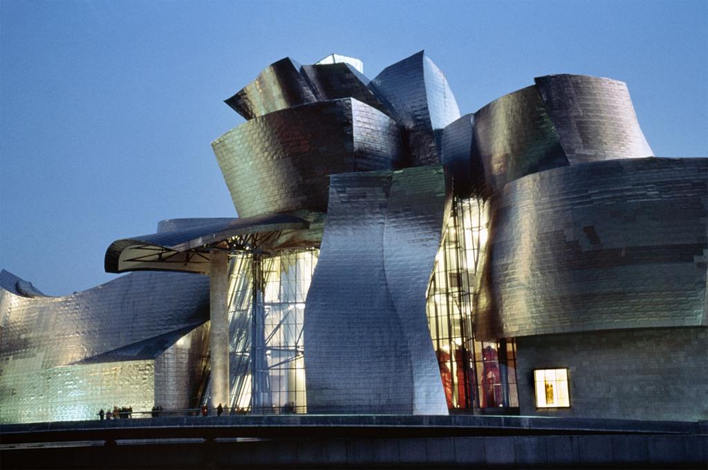 Architecture. Shiny metal tiles, glass, and stone shapes follow an asymmetrical, curvy building, several stories tall.
With not one, single, flat surface to be found anywhere on this highly unusual building, its undulating curves create a sense of movement. Large, circular shapes appear to be stacked unevenly upon each other and almost tipping to the side as the stories narrow toward the top of the building. Metal tiles cover Several of the stories, creating a highly reflective surface. This exterior image is from a clear day at dusk, so the surface reflects the deep blue of the sky. Lights illuminate the interior through tall, slightly curved and asymmetrical windows that reach several stories high. Human figures stand, silhouetted, in front of the building, giving a sense of its absolutely grand scale.

