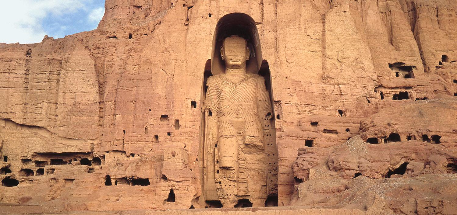 Sculpture. Nestled into a cliff face, a person stands wearing a sweeping robe.
The area around the person is barely larger than the person itself. The head is in tact but much of the face lost, so we only see the person’s closed lips and square jaw. The arms are down by the sides. There are more losses, especially below the elbows, but shallow lines suggest fabric draped across the shoulders and torso.
