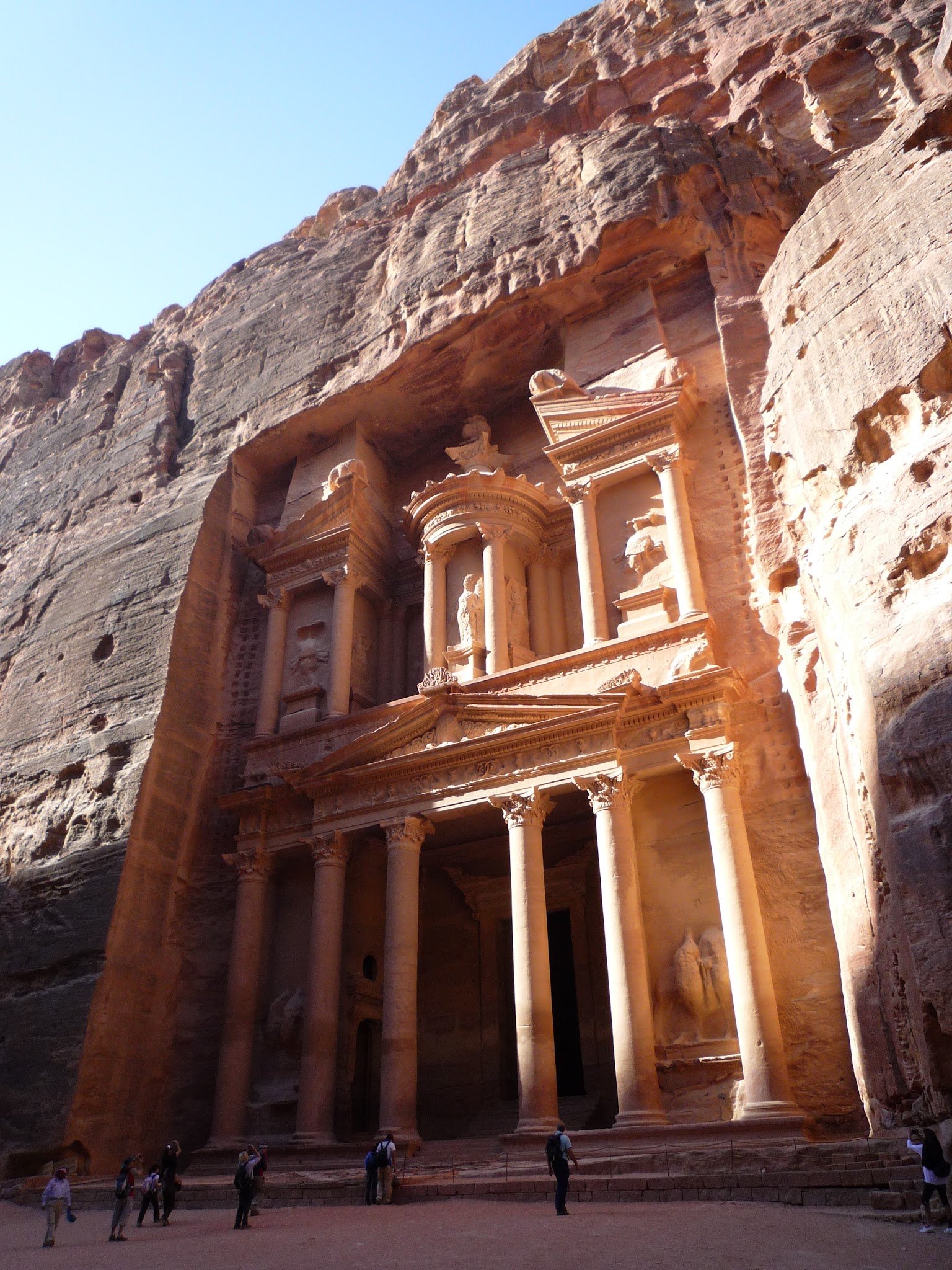Architecture. A two-leveled temple is cut directly into the surrounding cliffs.
The lower level has engaged columns flanking a rectangular opening. Above, three bays with shallow, rectangular niches are flanked by projecting columns, so there are deep gaps between each of the five elements. A rounded, tent-like canopy creates a little roof over the central bay. The bays to each side support the separated left and right halves of a pediment. An expanse of sand stretches in front of the structure, and the cliffs completely enclose the façade. A few people standing in front of the temple show that each level is at least four stories high.
