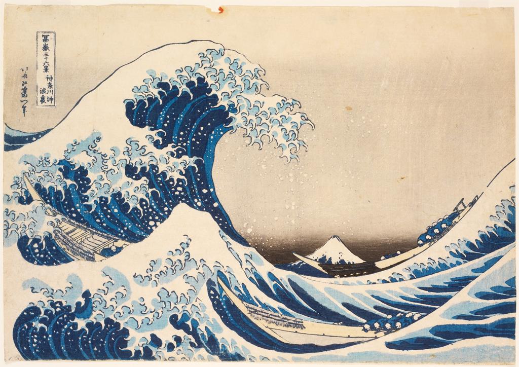 Color woodblock. Close to us, a vivid blue ocean wave rises and crests amid a froth of stylized white water.
The wave fills nearly the left half of the composition. The massive peak nearly breaks, its finger-like surf heavy along the back of the surge. Another peak slightly in front of it almost engulfs a long, narrow fishing boat. Another boat rides the neighboring swell, to our right. A mountain, tiny in the distance, is nestled in the troughs between the waves. The horizon is low, about a third of the way up the image. The sky around the mountain is smudged gray, and the sky above the parchment-white of the paper. A vertical cartouche and another column of Japanese characters are near the upper left corner.

