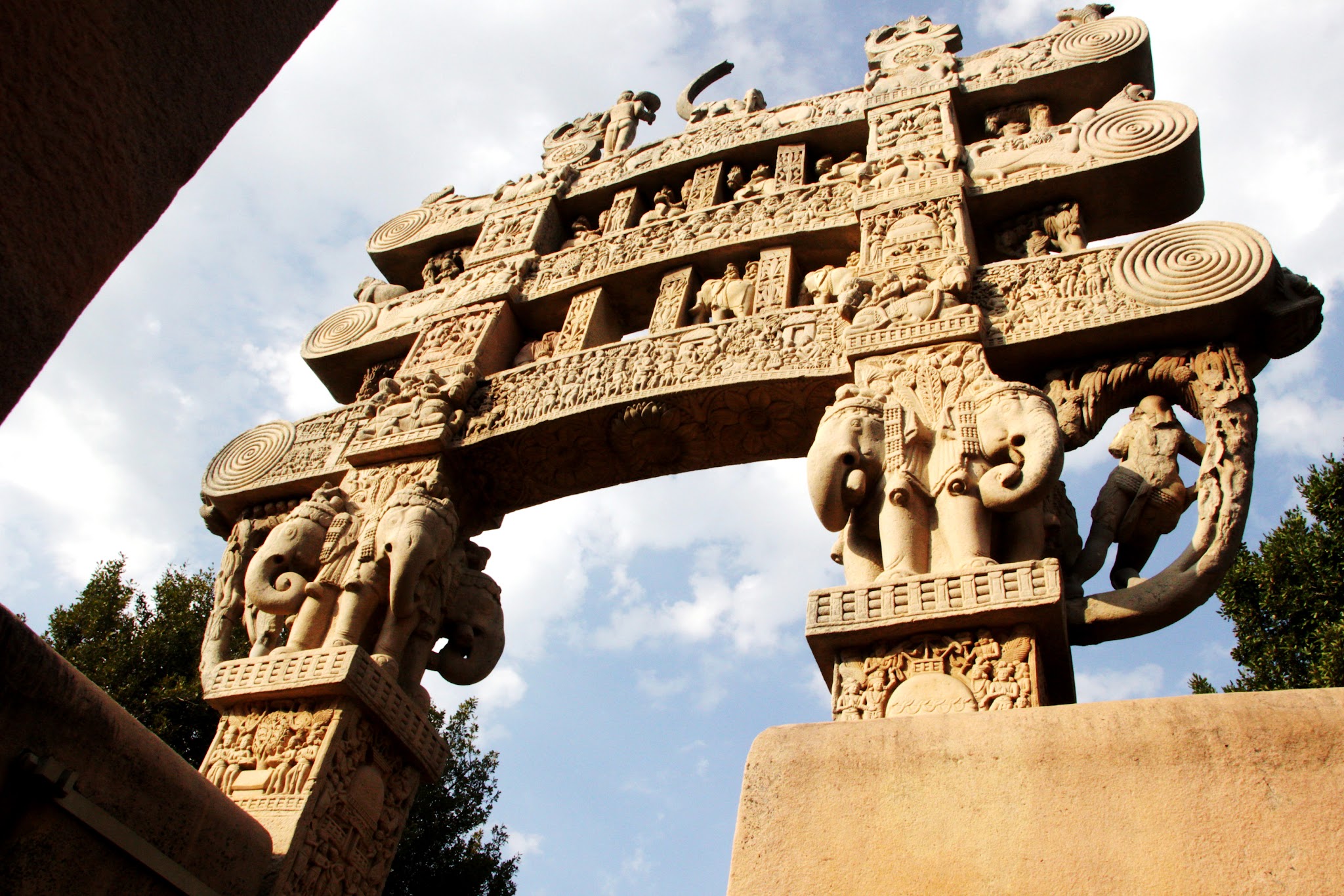 Architecture. Two stone pillars are woven through with three horizontal bands, all densely carved.
We look up sharply at capitals carved into the form of elephants. Above, three horizontal bands are supported, like stacked bridges, by the pillars. Much of the detail of the carvings cannot be made out in this image but there nested, concentric circles at both ends of the bands.

