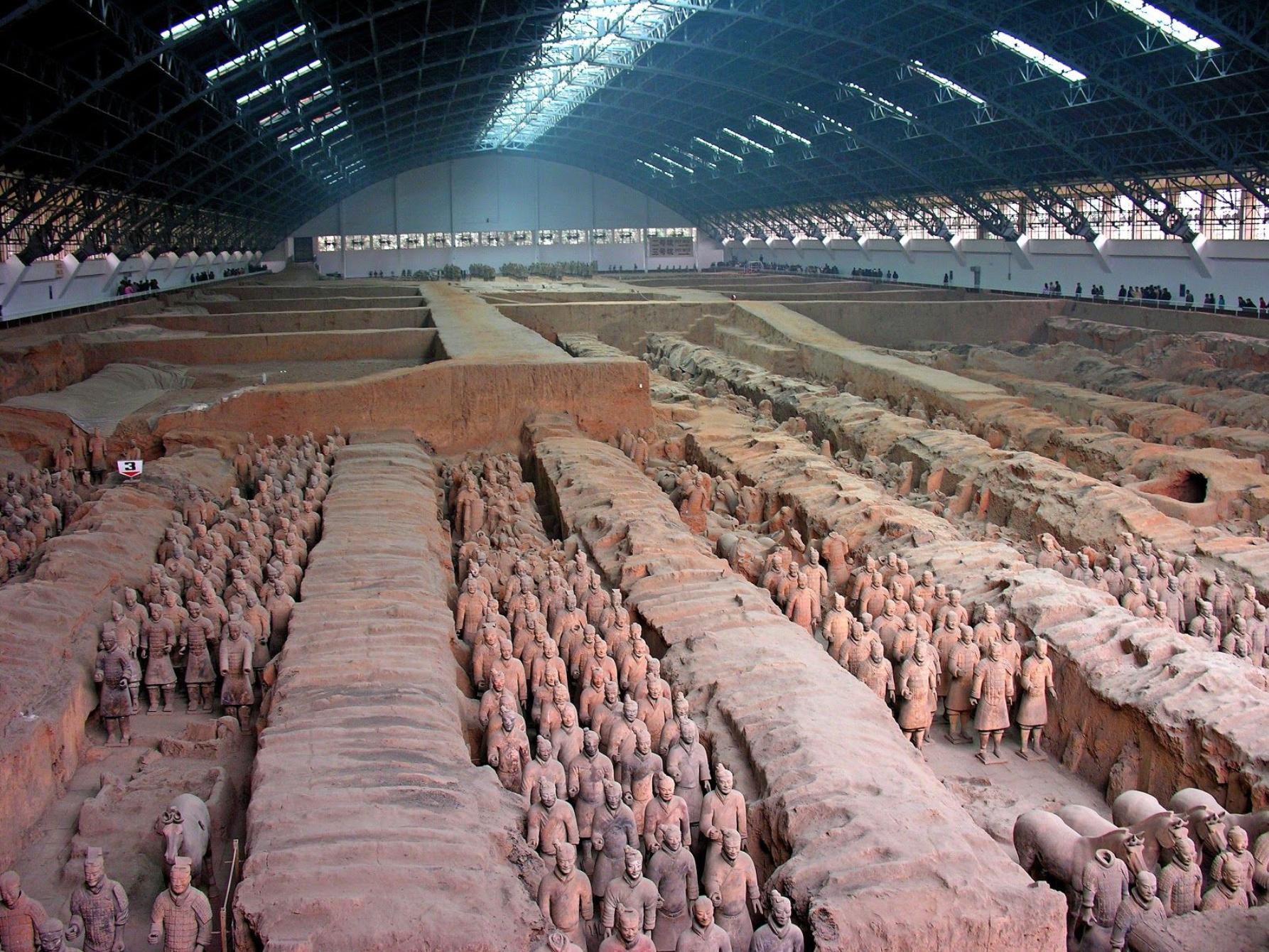  Sculptures. Hundreds of terracotta statues of men and a few horses fill a vast warehouse-sized building.
Separated by wall-like divisions, the statues are lined up in rows that stretch the length of the building. Closest to us, we can see that a few heads are missing. The people we can see stand squarely on both feet with their arms by their sides, though the exact position of their arms and hands varies.

