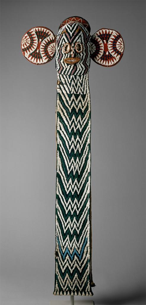 Mask. Wood, woven raffia, shells, beads, and cloth form a colorful mask with large ears and a long panel down the front.
The large, circular ears on either side of the mask give the appearance of an elephant, with the long panel down the front resembling a trunk. The eyes are close-set and wide open. The beads, cloth, shells, and raffia form dramatic patterns over every surface of the mask. The ears resemble the stitching patterns found on baseballs. The long panel down the front has darker and lighter lines zigzagging horizontally across, the face has similar lines, but vertically oriented. The dominant colors are reds, blues, and white, with subtle, gold accents on the face and ears.

