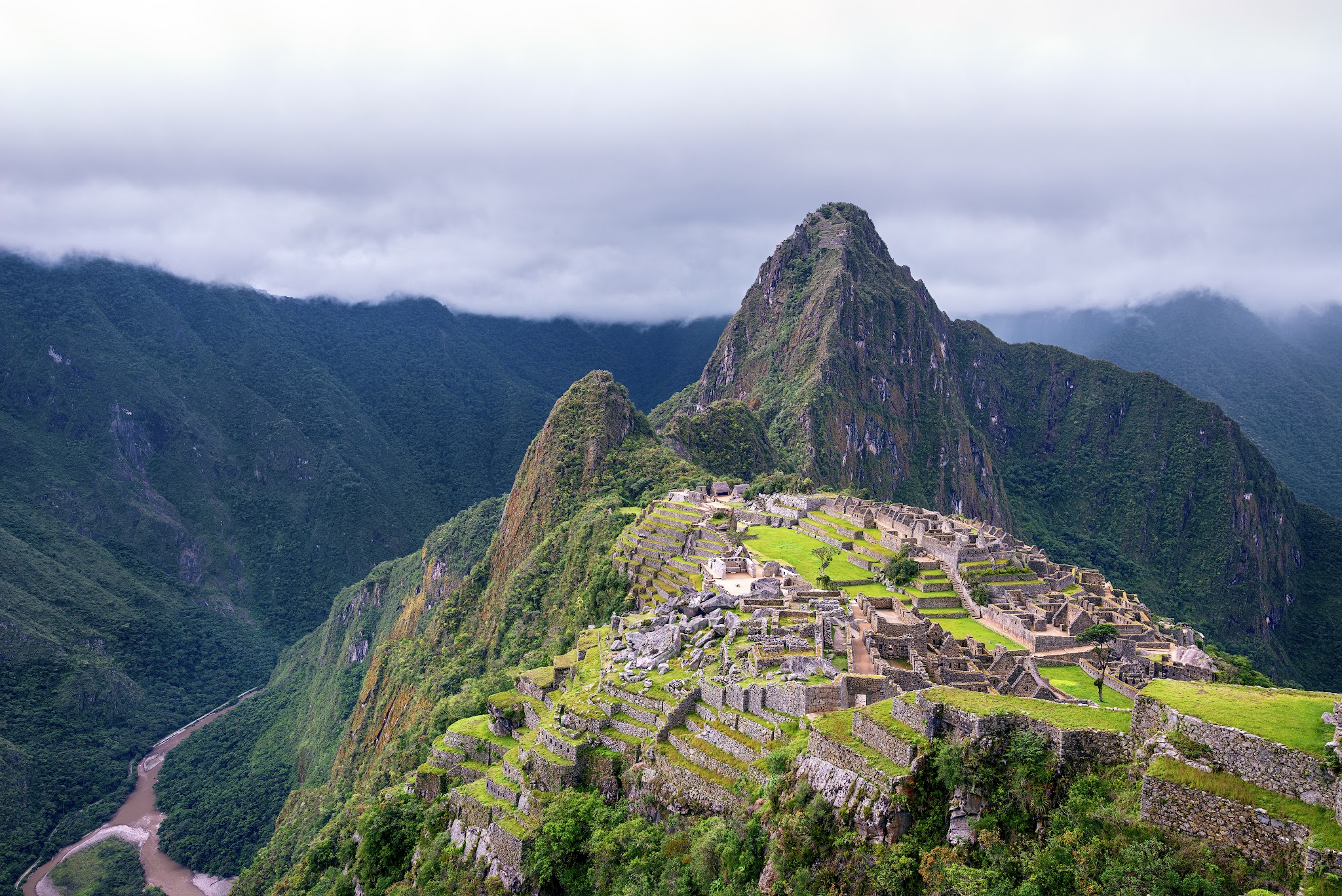 Architecture. Building complex sprawls across a mountaintop surface with terraced grass surfaces and many buildings.
This architectural complex appears to have hundreds of individual buildings, situated at the top of an isolated mountain. In the distance behind the built structures, another mountain range juts dramatically upwards towards a cloudy, stormy sky.

