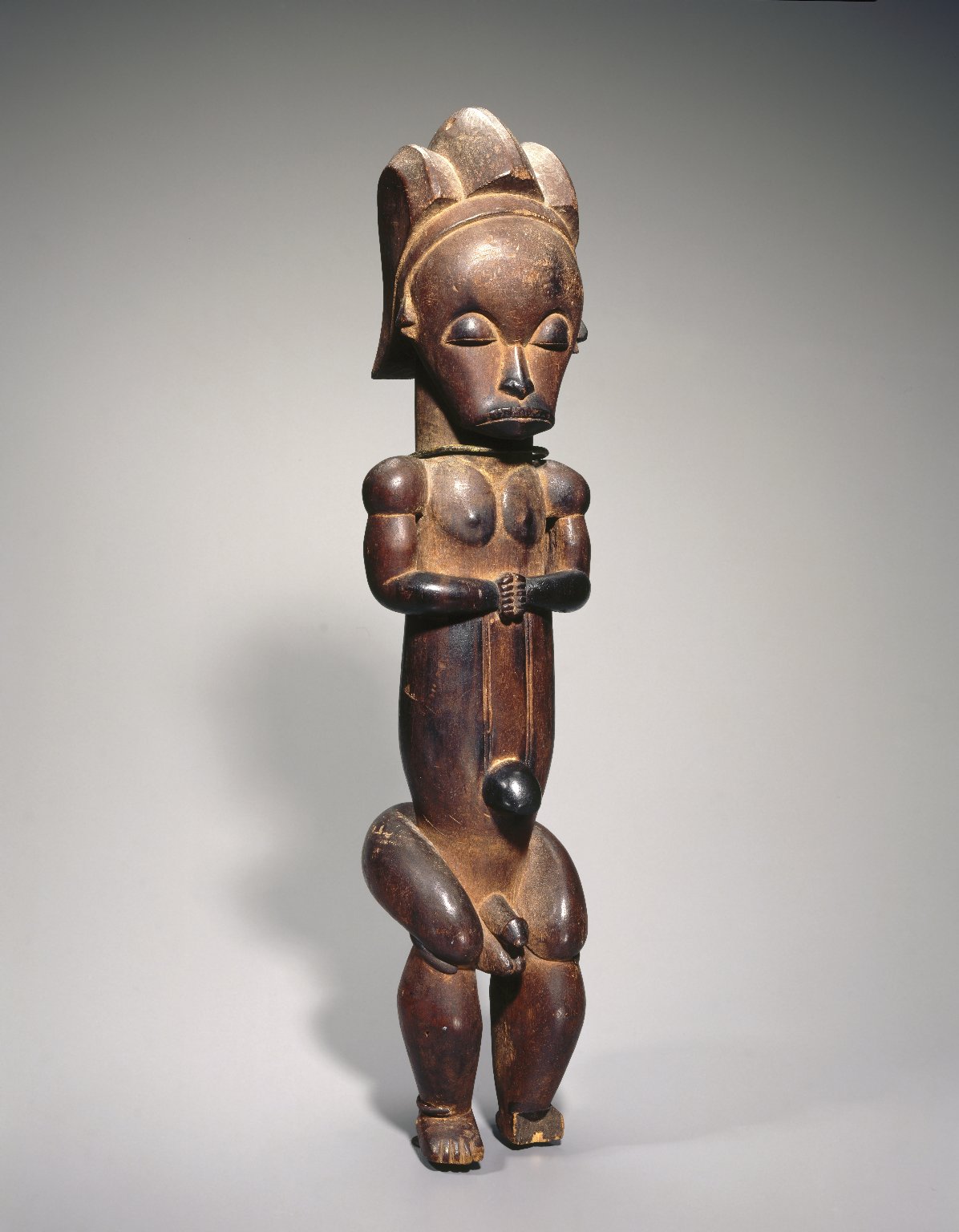 Sculpture. Long, narrow, wooden figure, with a relaxed expression and seated position, with disproportionate features.
This small statue is of a human figure in a relaxed posture. Its proportions are unusual, with an overly large head and an elongated torso. It appears to be seated, with a casual expression on its face and its hands clasped gently.

