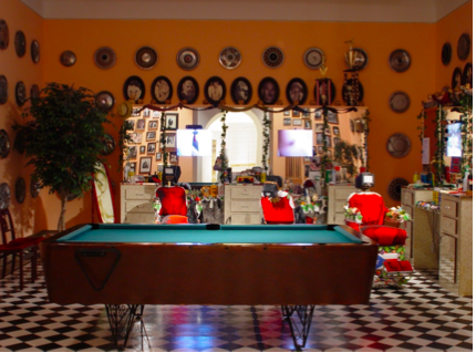Installation. Interior view of a kitschy barber shop with red chairs, a pool table, checkered floor, and framed photos.
At least twenty-five decorative pieces adorn the mustard-yellow walls in neat rows. Though they resemble ceramic ware, upon closer inspection they are actually hubcaps, interspersed with framed pictures of men's faces. Directly across the room from our vantage point is a large mirror, on top of which sit trophies and a hat. Under the mirror is a surface cluttered with various tools typical of a barbershop. There are three red chairs facing toward us as if waiting to welcome customers. Strapped to the top of each chair is a small video monitor. Reflected in the mirror are dozens of additional framed photographs, giving the sense an immersive experience.

