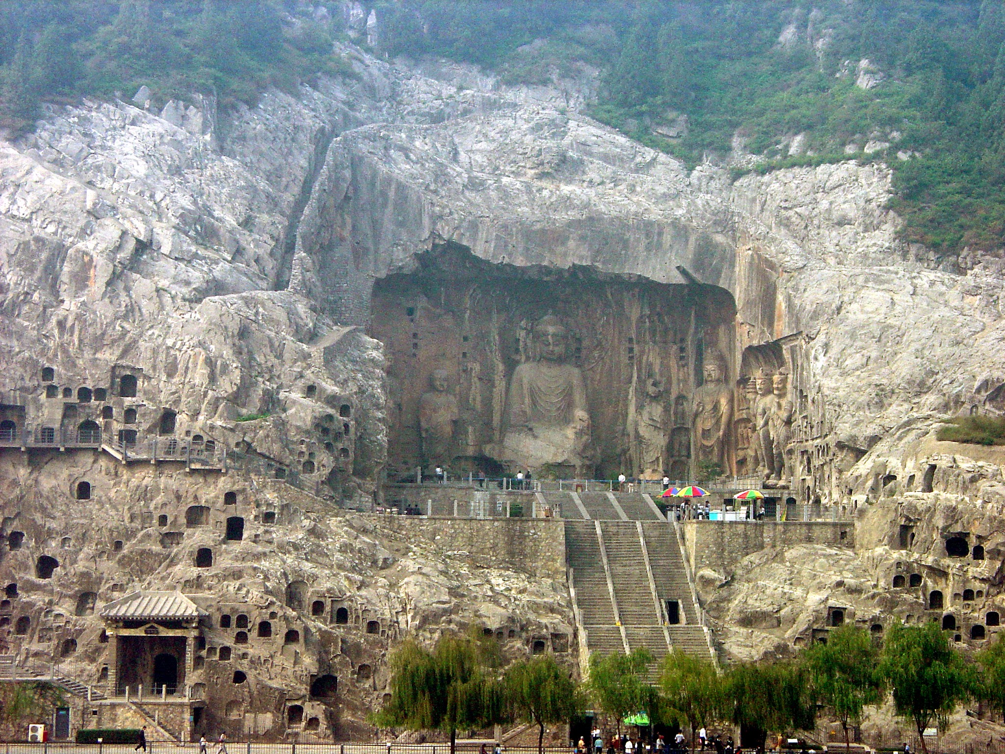 Architecture and sculpture. Statues and cave-like structures have been carved directly in the face of a sheer cliff.
A wide staircase leads up to a vast niche that encloses one seated person flanked by several others on a smaller scale. A couple of colorful patio umbrellas and miniscule people at the foot of the statues show that the central sculpture is several stories high. That person wears sweeping robes draped over crossed legs, and he appears to look down. Dozens of arched openings have been carved into the rock face around and below the statues.

