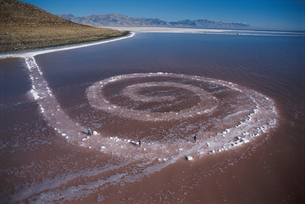Earthwork. A monumental, spiral-shaped artwork, made of rocks and earth, stretches out into a body of water.
This rocky structure stretches out across the water from a bright white shoreline. The scale is very large, a human could easily walk on top of this structure and follow its trajectory for several hundred feet. The structure resembles a rough, rocky pathway, about ten feet across, that extends from the shoreline out into the water in a straight line until it begins to curve, about a hundred feet in. At this point, the rocky path over the water forms a spiral pattern. The path of rocks winds inward in diminishing spirals until it ends at the center.

