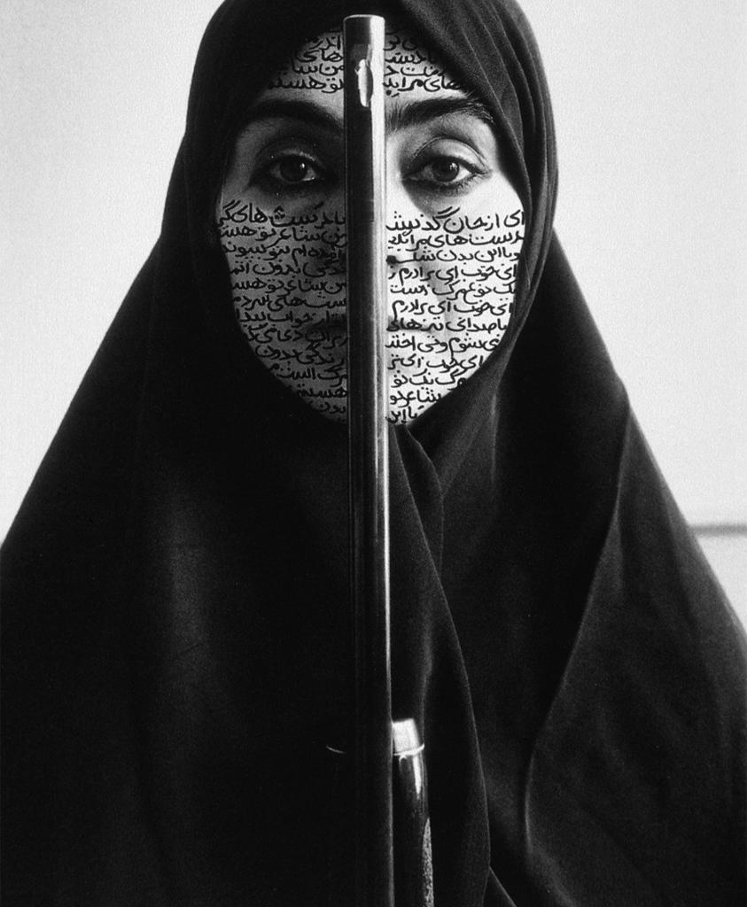 Photograph. A woman wears dark cloth over her head and shoulders, leaving her face, covered in Farsi writing, exposed.
This self-portrait is in black and white, the subject stares straight ahead at the viewer. The barrel of a rifle divides her face in half, casting a shadow on the left side. Dark eyes, brows, and eye makeup underscore the powerful contrasts in tone, and the neat rows of writing on her skin.

