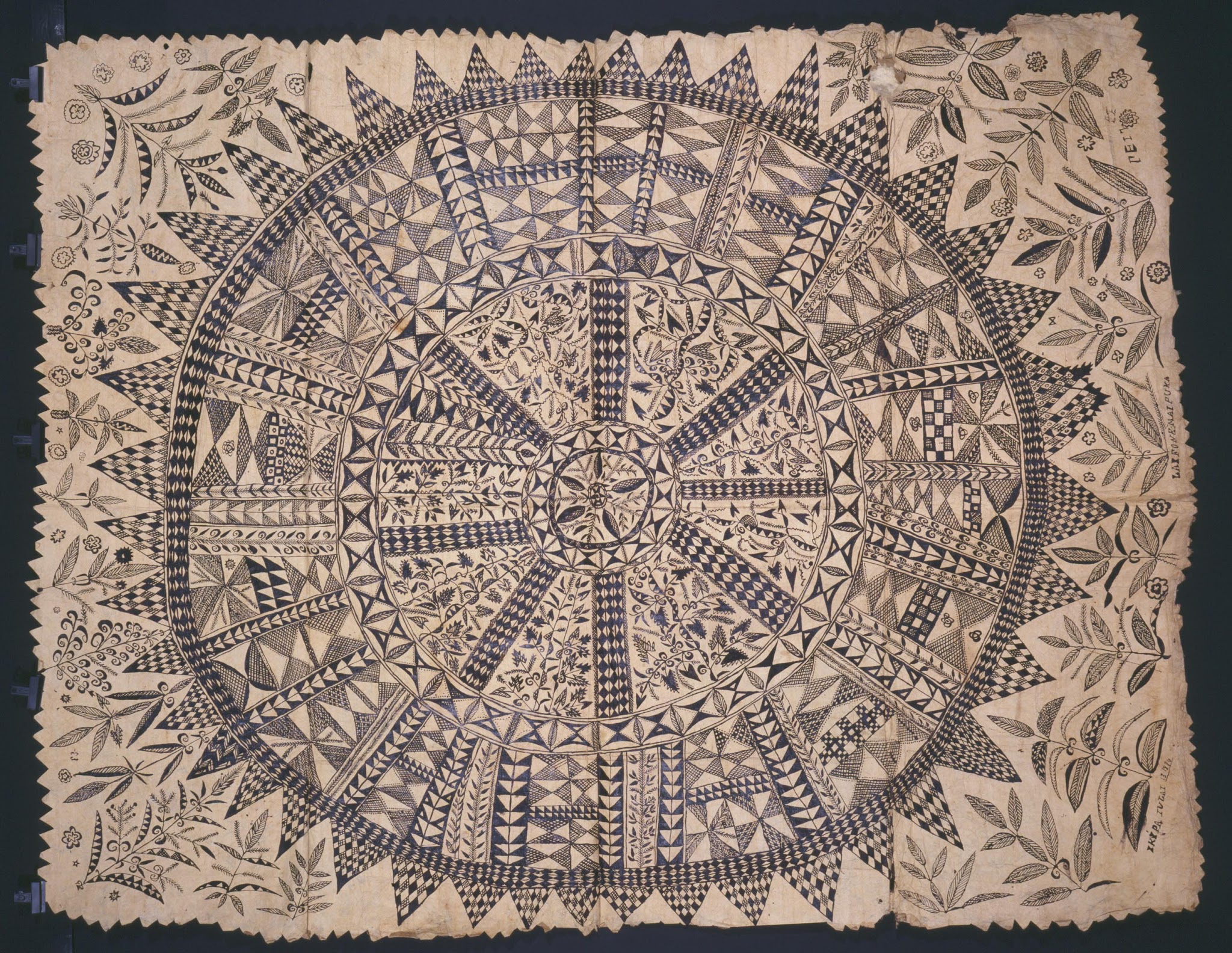 Textile. This hand-made cloth depicts a sunburst pattern of darker, concentric circles on a lighter background
There are three main circles to this design: one central, small circle with five spikes reaching outwards; the next a pattern with eight lines flowing from circle to circle, and the outermost circle a solid, dark line with spikes jutting outwards all around its circumference. The entire piece is adorned with similarly-proportioned, geometric decorations. including the cloth that extends outside of the outermost circle.

