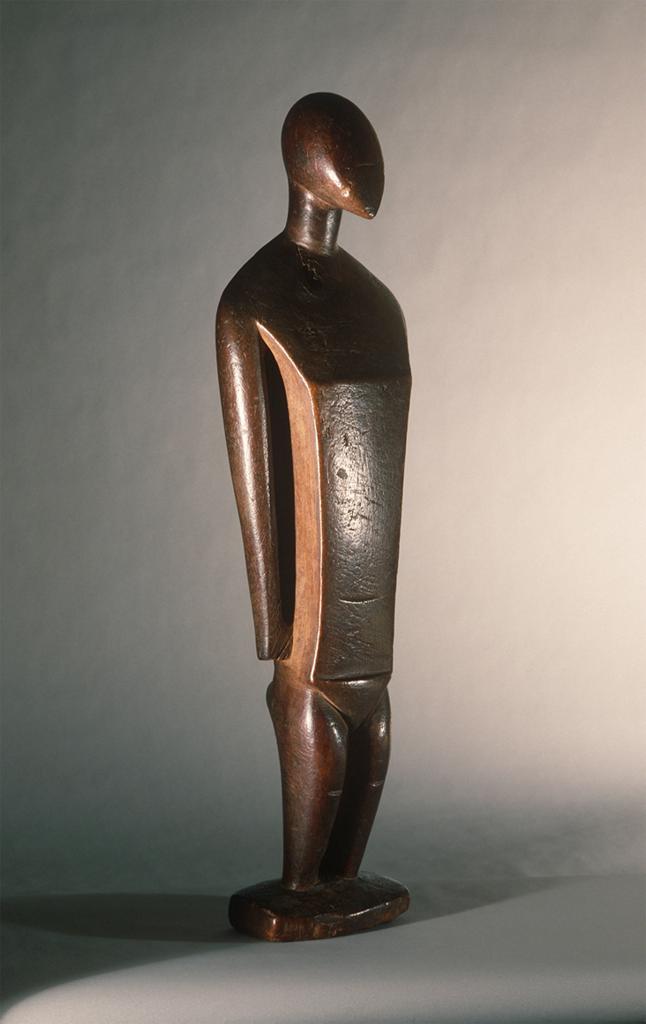 Sculpture. Human figure with a simple, geometric quality.The head is egg-shaped, the body long and narrow.
The egg-shaped head, on this abstracted sculpture of a human figure, comes to a point at the chin, which is slightly raised. A long neck descends into broad shoulders, with torso and arms of exaggerated length, the arms resting at the sides in a neutral position. The legs of the figure are shorter than the arms, giving the overall form a very tapered, stylized look.

