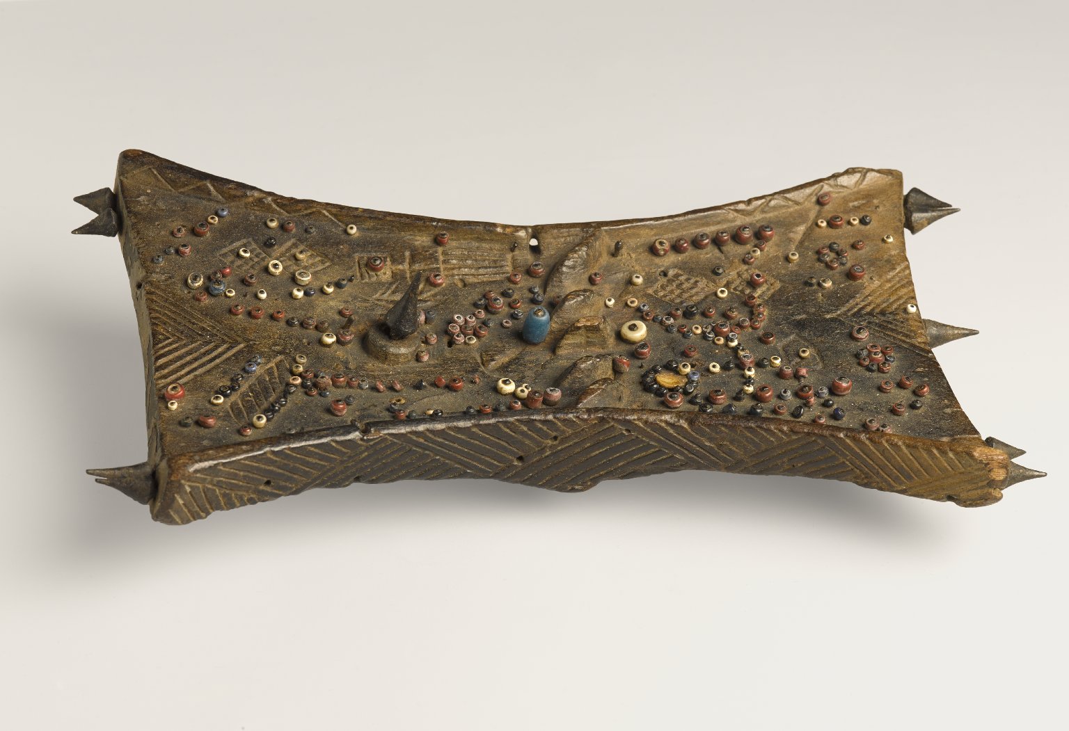 Sculpture. A board with multiple textures, in a bowtie shape, has images sculpted into it that appear to tell stories.
This sculpture contains wood, shells, metal, and beads. The small pieces of shells and beads are formed into several complex shapes, scattered across the main board. There are also low-relief incisions in the piece that describe unique patterns.

