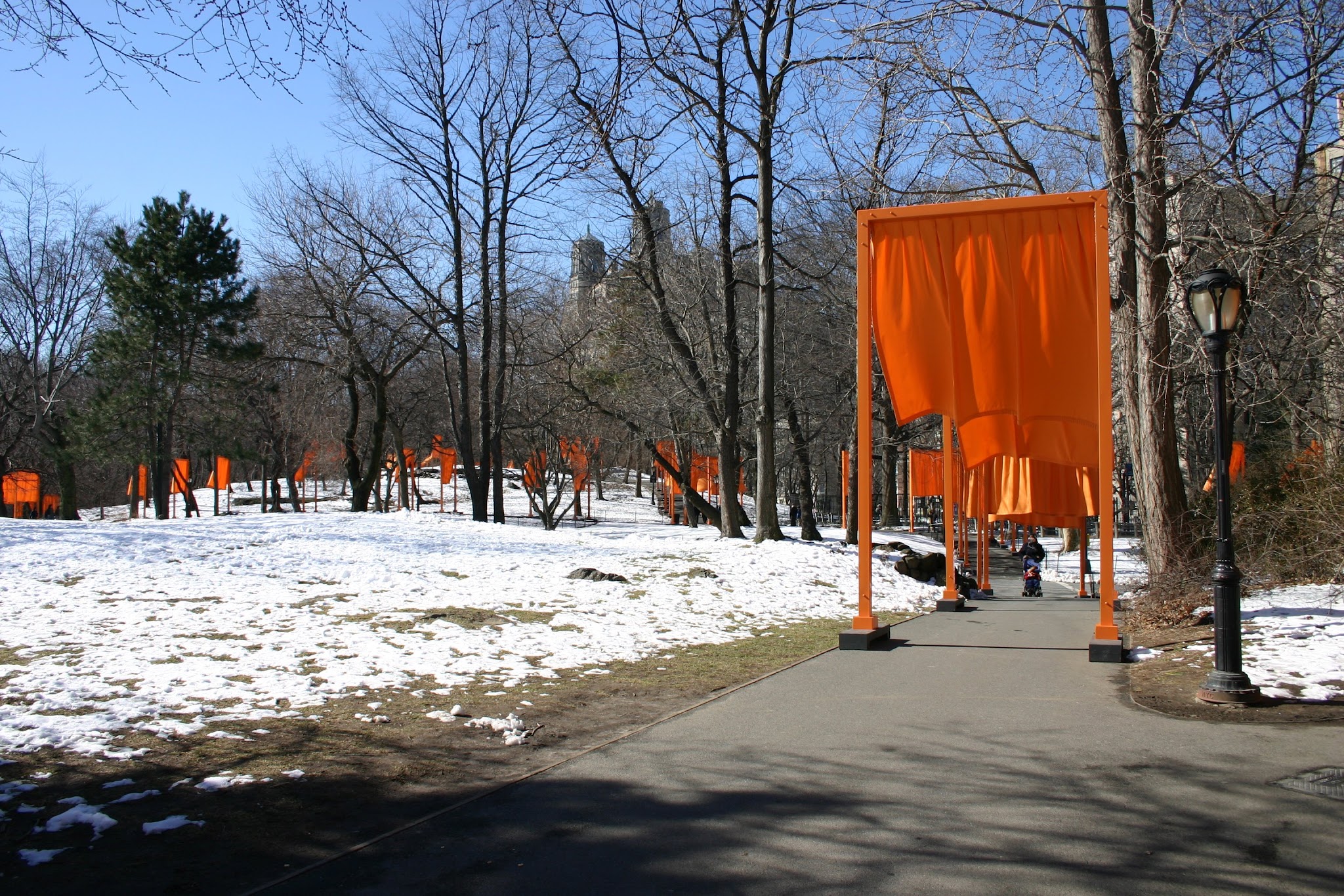 Installation. View of barren trees and a pathway with bright orange structures and tall buildings in the background.
The view of a park shows a walking paths, decorated with bright orange fabric. Each piece of fabric is supported by rectangular structures that allow it to hang and billow in the wind, tall enough for human beings to easily walk through.
