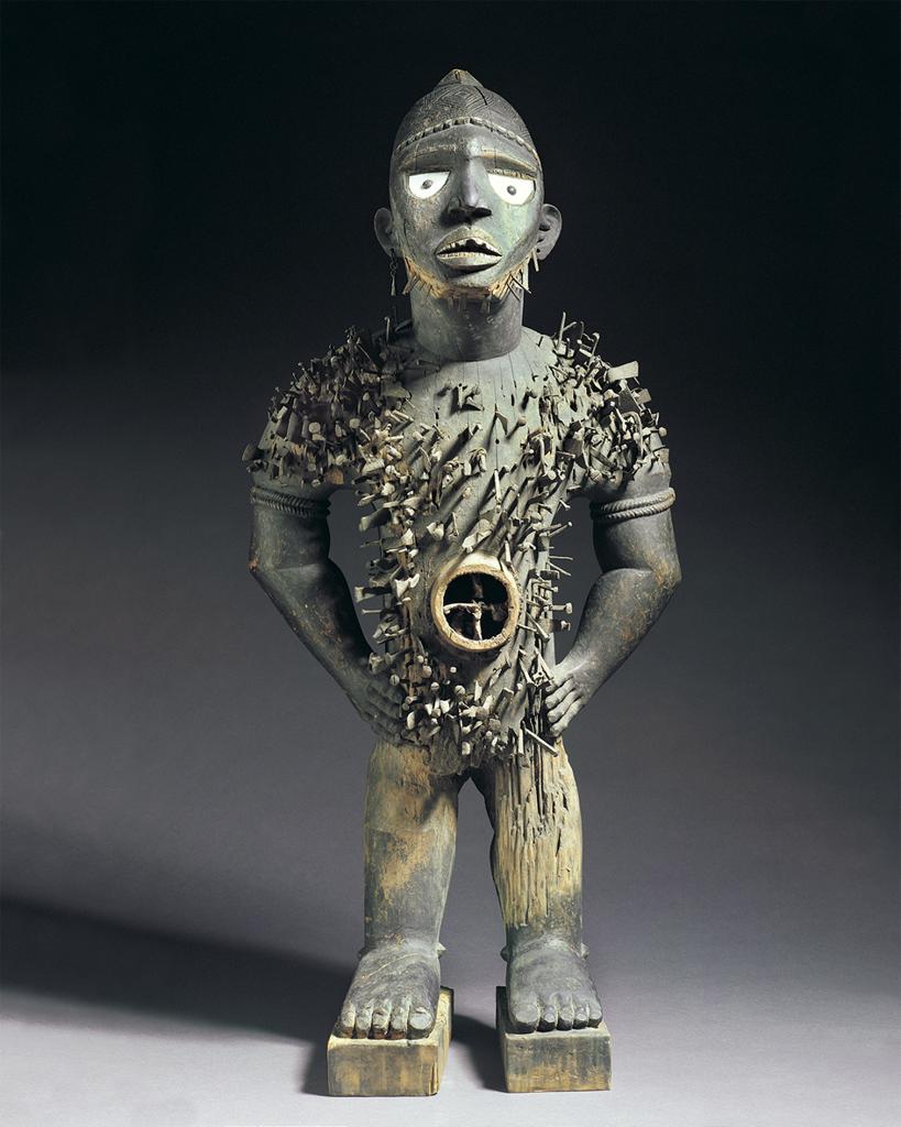 Sculpture. Frontal view of a small, wooden statue of a human figure, with intricately textured torso and wide eyes.
This small sculpture depicts a human figure whose head and legs are disproportionately large and small, respectively. Its bare feet are showing, its small legs in a strong stance, its hands on its hips. Hundreds of short, spiky objects pierce its torso, which has an empty, circular opening in the center.

