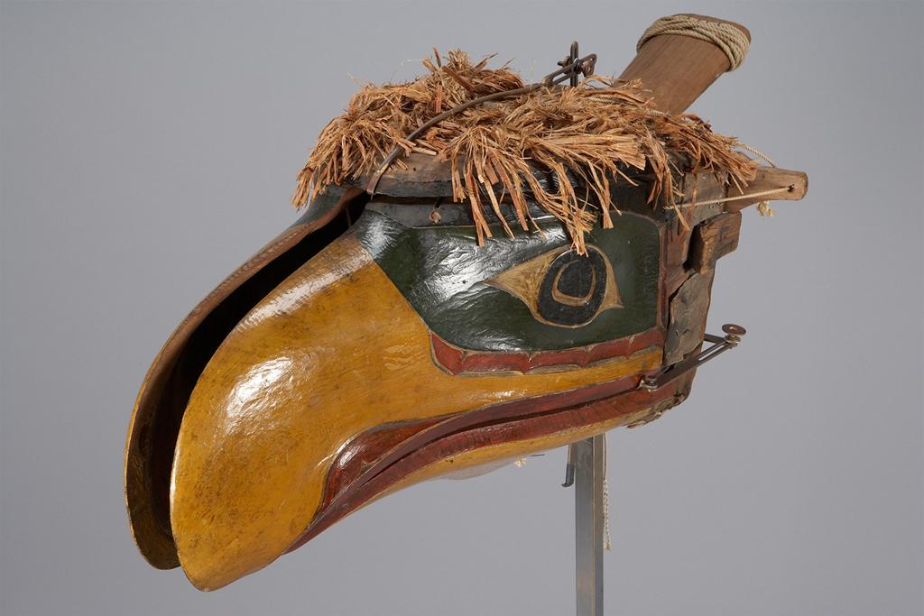 Carved mask. A wooden mask, painted in green, gold, and red, resembling the head of a bird, with thatched fibers on top.
This painted wooden mask is carved and painted to resemble a colorful bird. We see a three quarter view from the front, revealing a sharp, green eye, a golden beak, and a red-outlined beak opening. there is a split down the center of the beak, creating a narrow gap. The fibers decorating the top of the bird head resemble feathers, adding texture.

