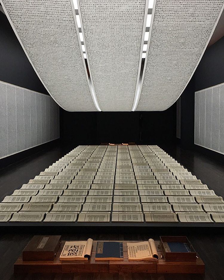 nstallation. A long, narrow, dark room, paper with writing is on the ceiling, walls, and in rows of books on the floor.
The dark walls of the room contrast with the lightweight and light-colored paper in three distinct locations. Three long scrolls of paper hang from the ceiling in an arc that recedes towards the far wall, and lights on the ceiling illuminate the paper from behind. The paper contains neat, tiny, lines of shapes that resemble Chinese characters, but are not legible. The two side walls also hold long scrolls of paper, but these are each flatly mounted to the wall. They also contain similar writing, broken up into multiple columns of uniform height and width. On the floor are rows of open, hand-bound books, also with the same characters. The curves of the open books mimic the curve of the scrolls hanging from the ceiling, giving a soft, airy appearance. In the foreground, closest to the viewer, there is a wooden fixture on the ground, which holds three closed books and two open books.
