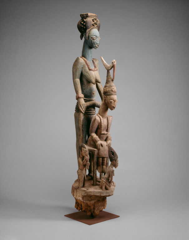 Sculpture. A sculptural group has four figures: one tall woman, one seated man, and two smaller figures at their feet.
This sculpture is a detailed, somewhat stylized and abstracted, depiction of a woman of impressive stature, wearing an elaborate headdress, standing protectively behind a seated man with an even more elaborate headdress, which has a bird-like figure perched on top. On the ground, at their feet, are two small figures with similar dress and headwear. One is kneeling, one is crouching slightly.

