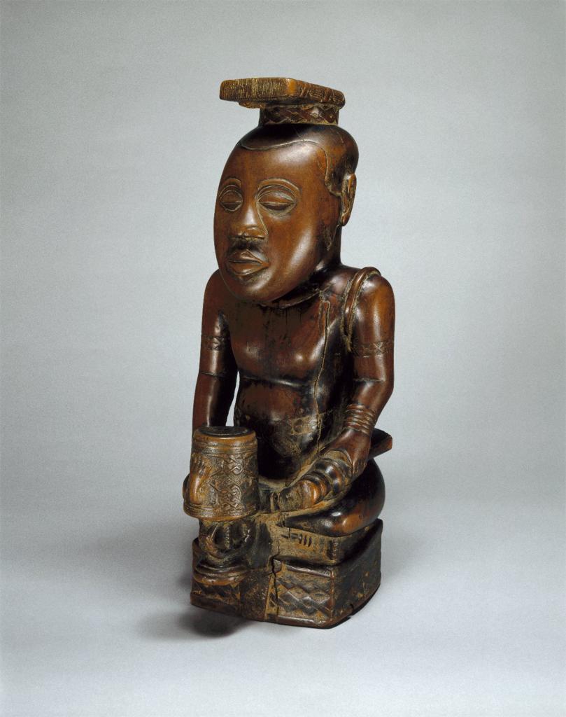 Sculpture. Small statue of human with exaggerated facial features and proportions, cross-legged and holding a drum.
This small statue depicts a kneeling man holding a drum. The bald head is exaggerated in size to the point that it is as large as the torso of the figure. Its eyes and mouth are closed, relaxed. The skin of the bare torso is carved with symbols at the upper arms and bracelets at the wrists. On its head is a tight-fitting, crown-like headdress with a flat, rectangular slab on top. Its left hand is missing, the arm ending abruptly at the wrist. On the drum is a symbol of a severed hand.

