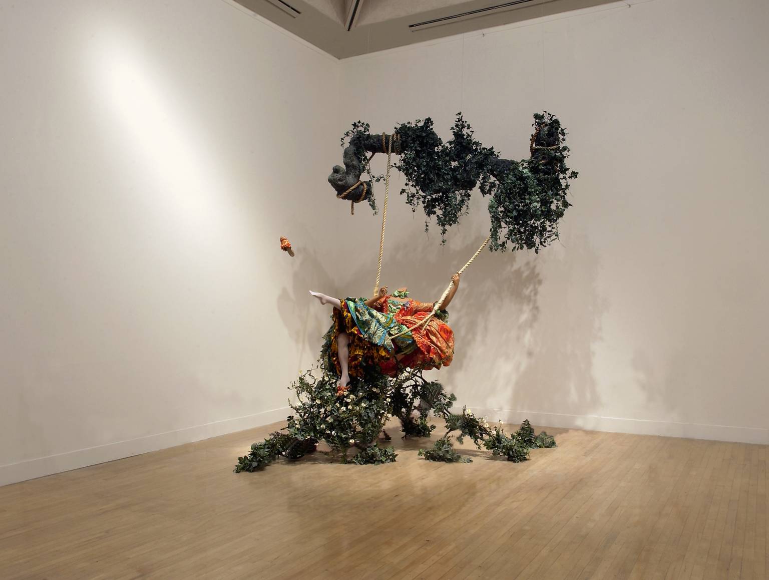 Sculpture. A life-sized mannequin, suspended from the ceiling on a swing, wears brightly-colored fabrics.
Above and below this figure are splashes of greenery, as if the subject is swinging through a lush garden. It is clear, however, that the scene is set within an interior space with white walls.

