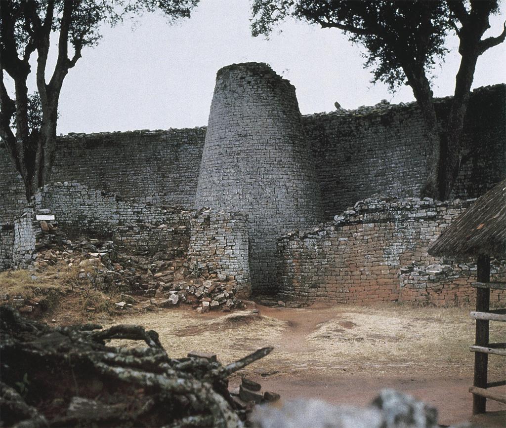 Architecture. A tall, cone-shaped structure, with imposing walls stretching out either side, made of stone masonry.
This enclosure has a multitude of individual pieces in its stone masonry. There is a central, cone-shaped structure that rises high above the walls extending outwards from its sides. It appears that additional structures might once have been part of this section of a massive wall, but they are clearly ruins now.

