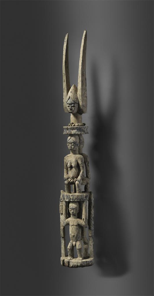 Sculpture. Small, narrow figure with three different levels of human/humanoid figures. Two pointed shapes at the top.
This small statue depicts three separate, human figures on top of one another. At the base is a nude, boyish figure. Above this is a seated, more feminine looking figure, also nude. At the top is a truncated figure with a calm, closed-eye expression.

